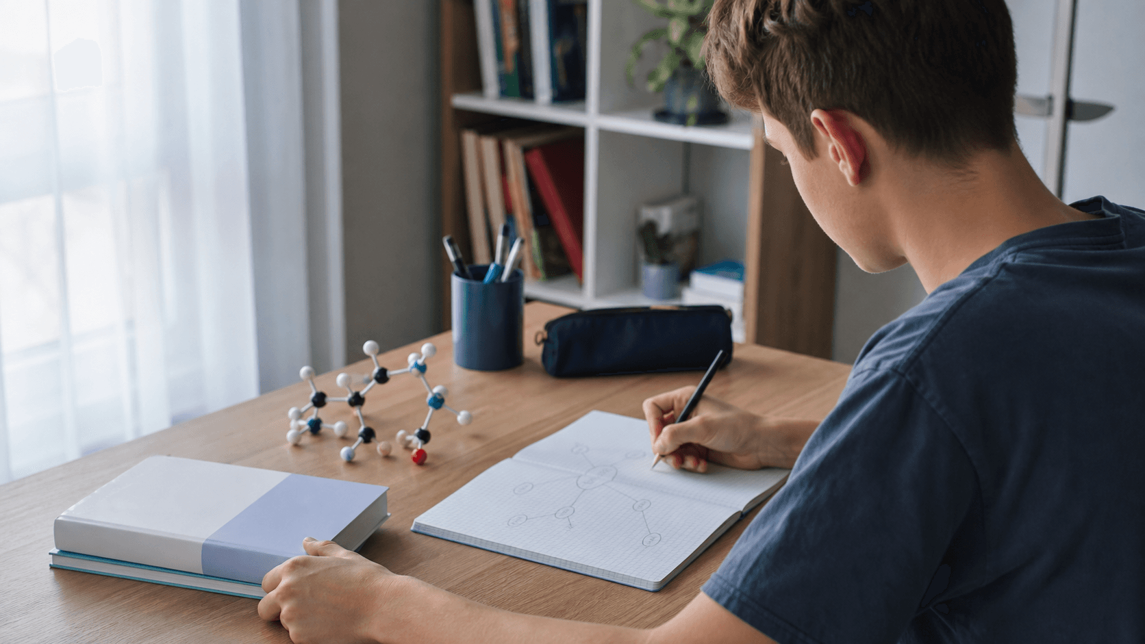 Elev de liceu văzut din spate, studiind chimia la birou cu un caiet, un model molecular și manuale