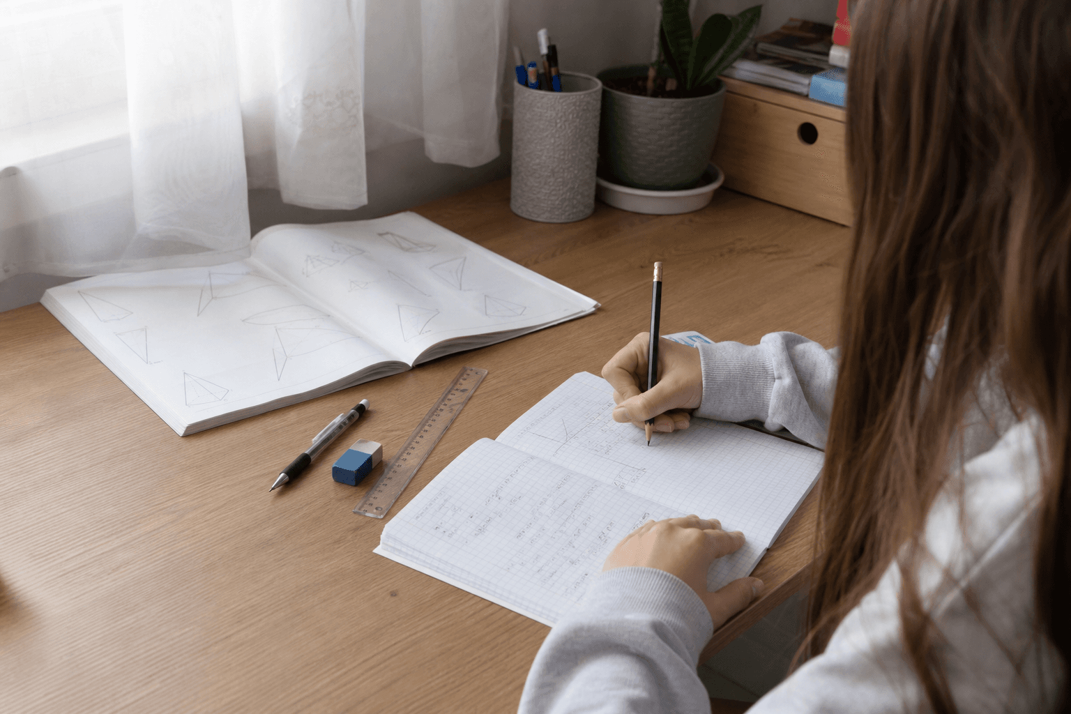 Elevă văzută din spate, rezolvând exerciții de matematică într-un caiet dictando, la un birou luminat natural
