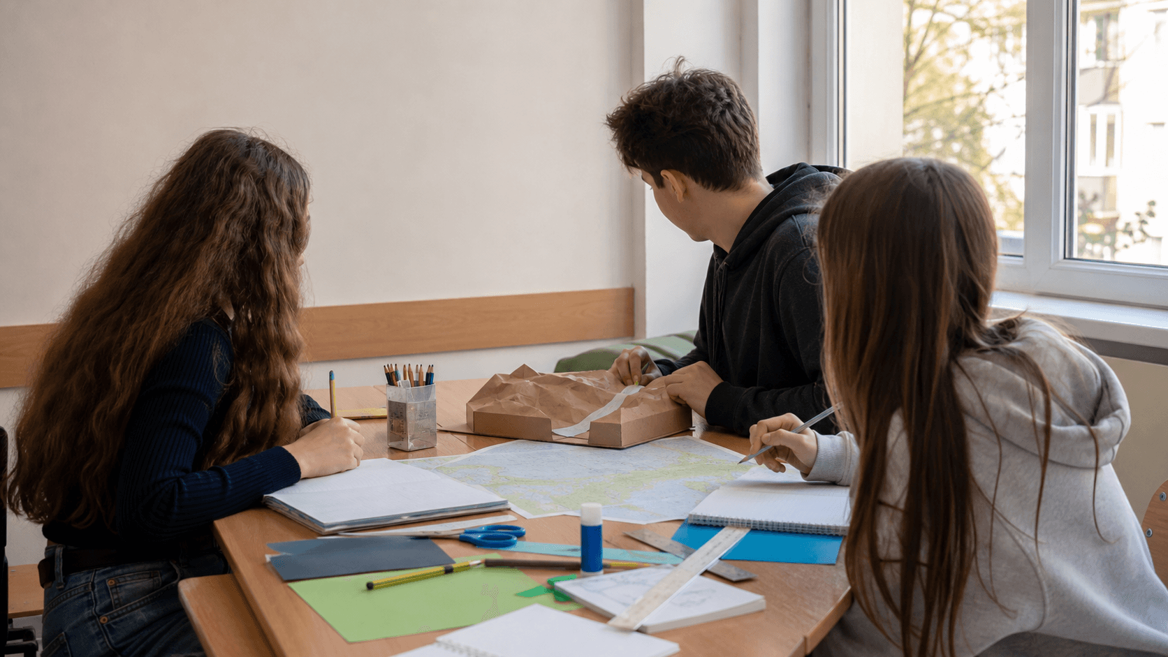 Elevi care pregătesc un proiect școlar pe o masă, cu caiete, imagini și materiale organizate